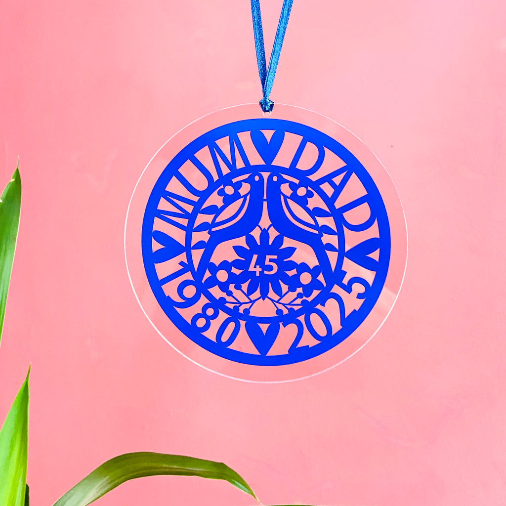 Sapphire blue acrylic anniversary decoration featuring “Mum & Dad,” two lovebirds, floral details, and the years 1980–2025 with “45” in the center. This elegant cut-out ornament hangs on a blue ribbon against a soft pink background, making it a perfect 45th wedding anniversary keepsake or gift.