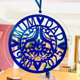Beautiful sapphire blue acrylic hanging ornament celebrating a 45th wedding anniversary. Features "Mum & Dad," two lovebirds, floral design, and the years 1980–2025 in an elegant cut-out style. Shown hanging in a bright home interior, perfect as a keepsake or window decoration for a special milestone.